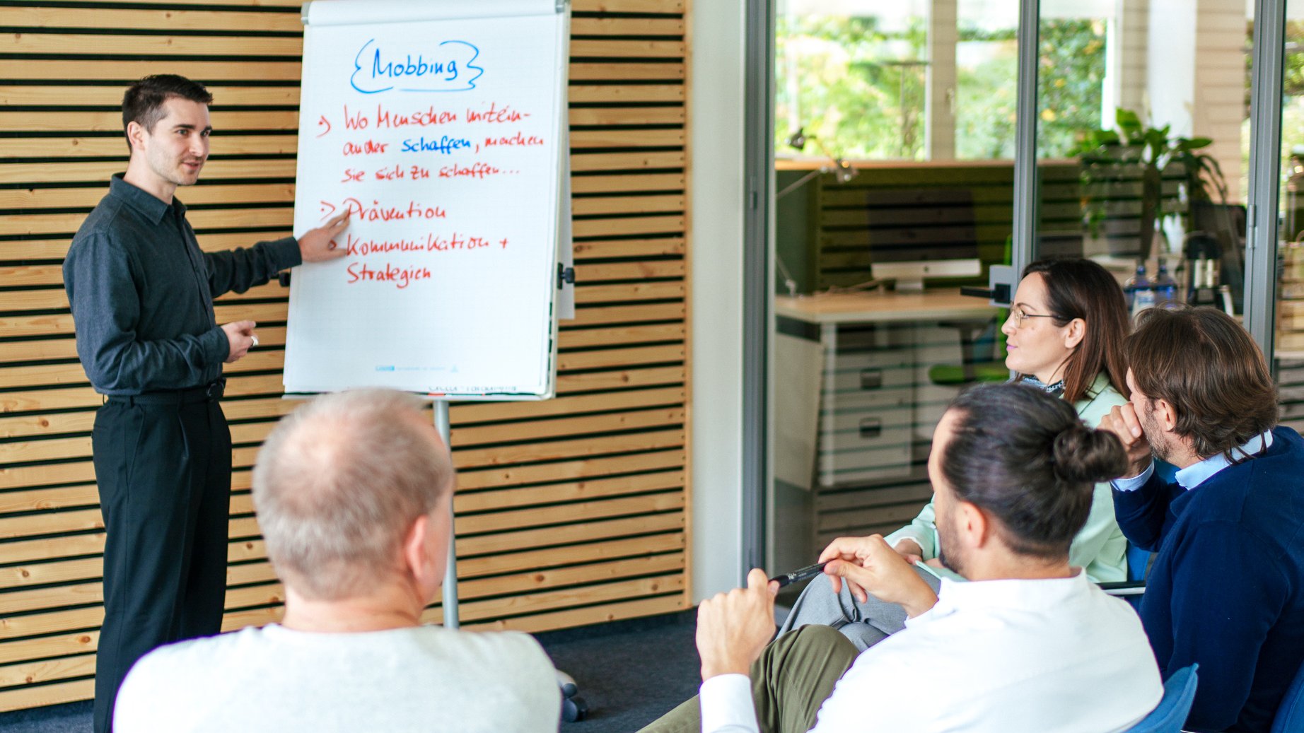 Vincenzo Rocco steht vor einer kleinen Gruppe sitzender Personen und präsentiert Inhalte zum Thema Mobbing auf einem Flipchart