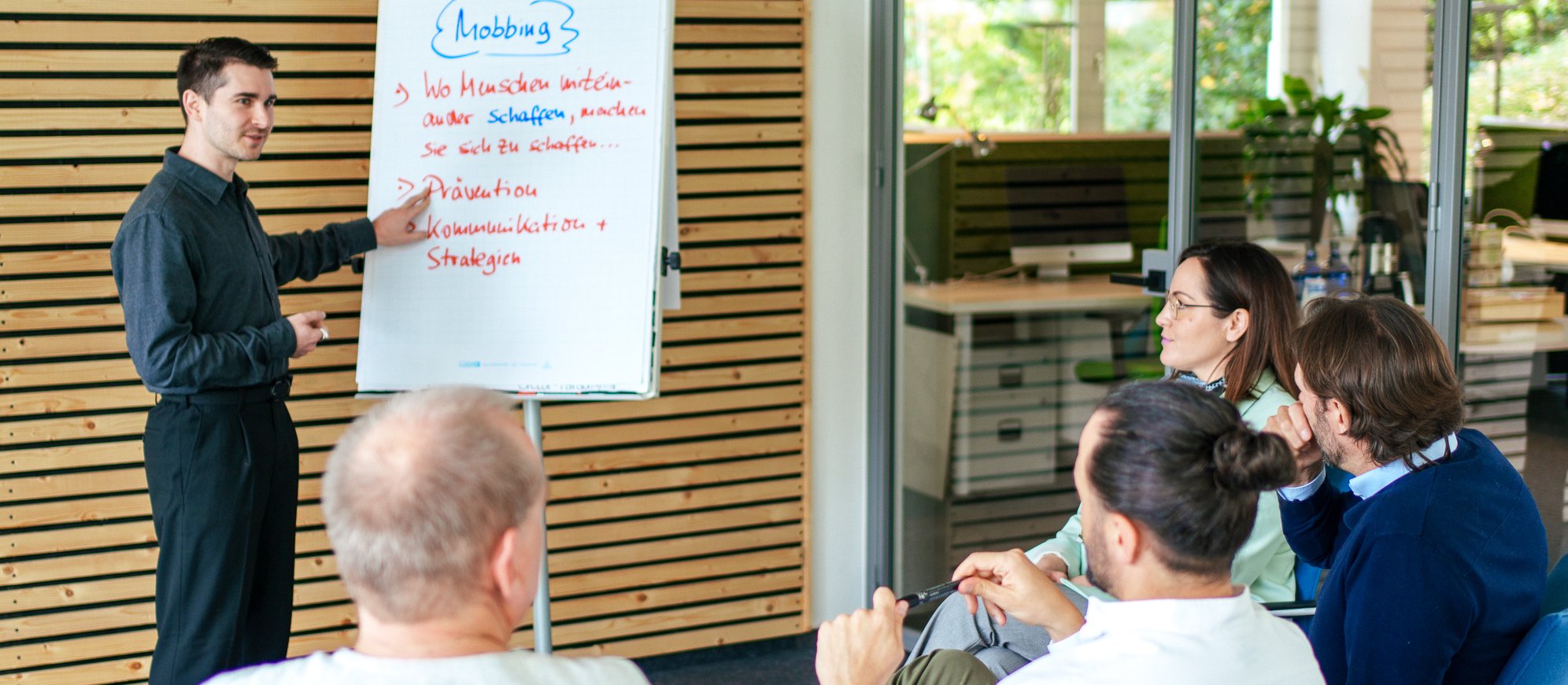 Vincenzo Rocco steht vor einer kleinen Gruppe sitzender Personen und präsentiert Inhalte zum Thema Mobbing auf einem Flipchart