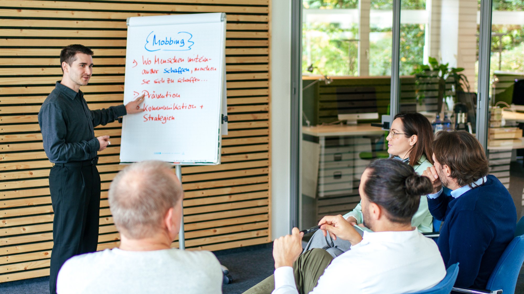 Vincenzo Rocco steht vor einer kleinen Gruppe sitzender Personen und präsentiert Inhalte zum Thema Mobbing auf einem Flipchart
